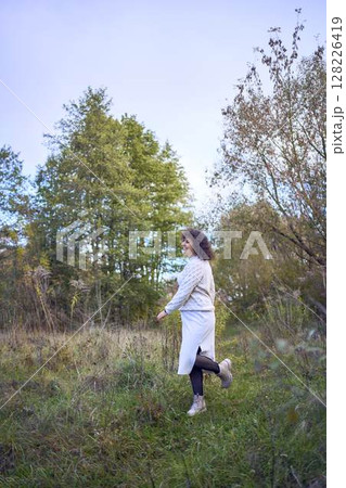 middle-aged woman with dark curly hair in light beige suit enjoying nature in autumn forest 128226419