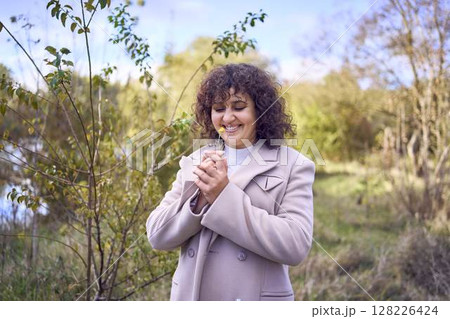 middle-aged woman with dark curly hair in light beige suit and coat enjoying nature in autumn forest 128226424