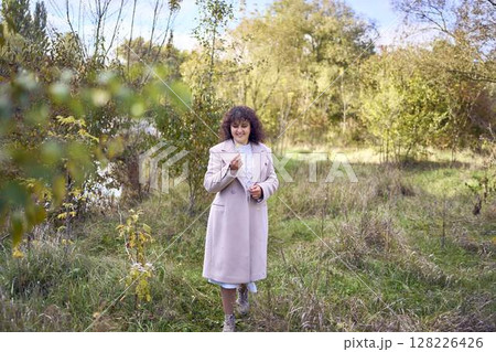 middle-aged woman with dark curly hair in light beige suit and coat enjoying nature in autumn forest 128226426