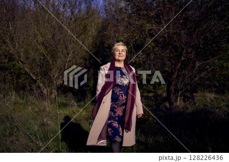 elderly woman with gray hair in light beige coat enjoying nature in autumn forest 128226436