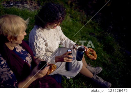 middle-aged daughter and old mother eating pastries and drinking tea from thermos cups on a picnic in the autumn forest 128226473
