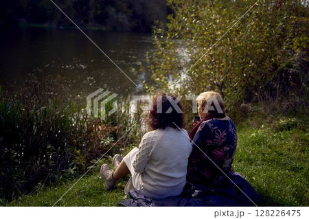 middle-aged daughter and old mother eating pastries and drinking tea from thermos cups on a picnic in the autumn forest middle-aged daughter and old mother eating pastries and drinking tea from thermos cups on a picnic in the autumn forest 128226475