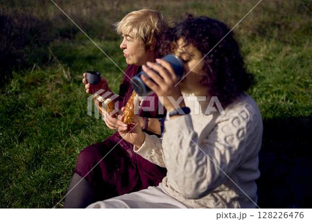 middle-aged daughter and old mother eating pastries and drinking tea from thermos cups on a picnic in the autumn forest middle-aged daughter and old mother eating pastries and drinking tea from thermos cups on a picnic in the autumn forest 128226476
