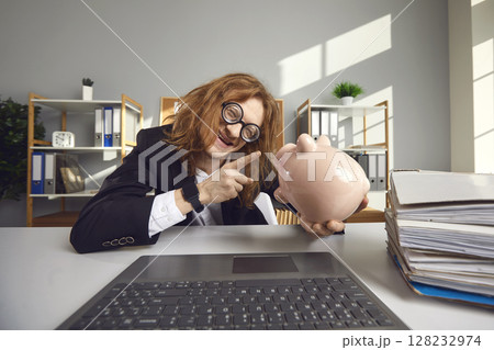 Strange crazy guy freak talks to a piggy bank sitting at a table in front of a laptop in the office. Strange crazy guy freak talks to a piggy bank sitting at a table in front of a laptop in the office. 128232974