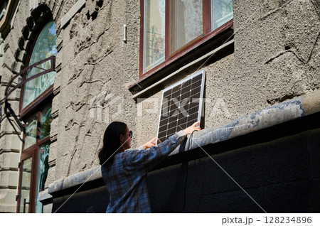 Woman holding photovoltaic solar panel in front of historical building, wearing plaid shirt and sunglasses. Concept of integration of sustainable renewable energy sources into old architecture. Woman holding photovoltaic solar panel in front of historical building, wearing plaid shirt and sunglasses. Concept of integration of sustainable renewable energy sources into old architecture. 128234896