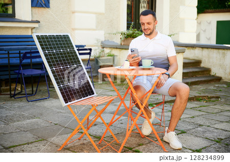Young man charging smartphone from solar panel while sitting in cafe. Handsome male drinking coffee and checking his phone next to solar battery. Conscious male choosing sustainable lifestyle. Young man charging smartphone from solar panel while sitting in cafe. Handsome male drinking coffee and checking his phone next to solar battery. Conscious male choosing sustainable lifestyle. 128234899