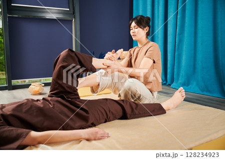 Masseuse making thai yoga massage. Woman therapist doing traditional massage treatment for female patient indoors. Client lying on mat, meditating, enjoying hand massage in calm atmosphere. 128234923