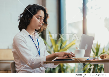 Young woman typing on laptop during lunch break in bright office cafeteria with coffee Young woman typing on laptop during lunch break in bright office cafeteria with coffee 128234957