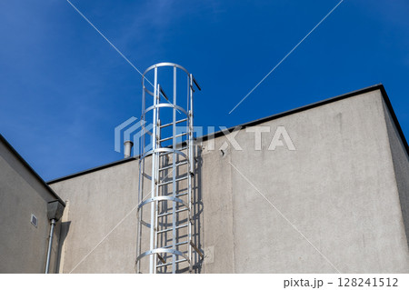 Tall silver industrial caged access ladder firmly attached to a plain grey concrete building wall, representing safety, emergency exit, and rooftop access solutions 128241512