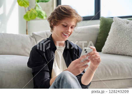 Happy joyful handsome teenager guy using smartphone sitting on floor at home. Happy joyful handsome teenager guy using smartphone sitting on floor at home. 128243209