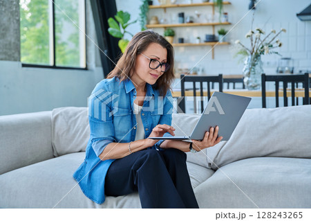 Portrait of middle aged woman using laptop sitting on sofa at home 128243265