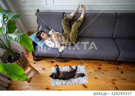 Bored woman with feet up on couch in smartphone, browsing internet chats and social feed near cat 128243317