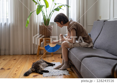 Caring woman pet owner gently fanning cat curling on floor to ease heat stress at home, no AC 128243516