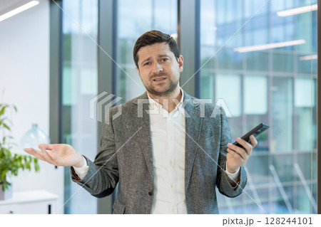 Portrait of a worried young businessman standing in a suit in the office, spreading his arms and looking at the camera sadly. Portrait of a worried young businessman standing in a suit in the office, spreading his arms and looking at the camera sadly. 128244101