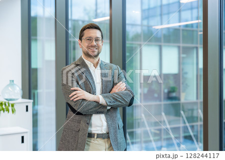 A smiling young man, founder and owner of a firm and company, stands in the lobby of an office building, arms crossed on his chest and confidently looking into the camera. 128244117