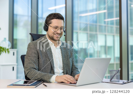 Smiling young office worker in headset sitting at desk in office and working on laptop. Smiling young office worker in headset sitting at desk in office and working on laptop. 128244120