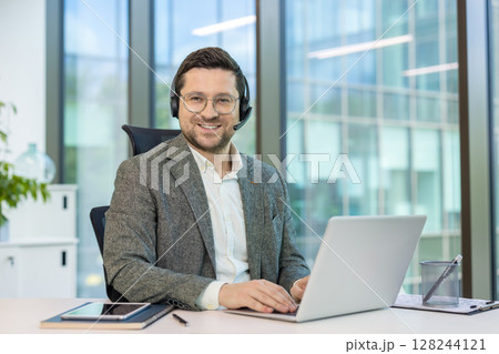 Portrait of a smiling young man in a suit and headset sitting at a working office desk and looking at the camera. Portrait of a smiling young man in a suit and headset sitting at a working office desk and looking at the camera. 128244121