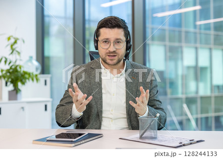Portrait of a young man in a headset and computer sitting in an office at a table and holding an online video conference, explaining by gesturing with his hands. 128244133