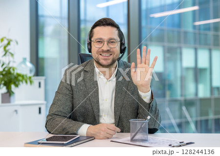 Portrait of a businessman in a headset and suit sitting at a desk in the office, smiling and waving at the camera. 128244136