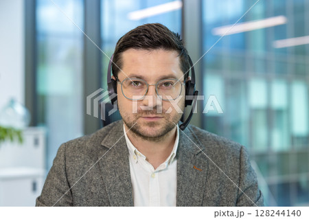 Close-up portrait of a young serious male office worker and assistant sitting at his workplace in a headset and looking intently at the camera. Close-up portrait of a young serious male office worker and assistant sitting at his workplace in a headset and looking intently at the camera. 128244140