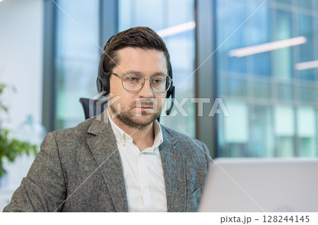 Close-up photo of a young man in a suit and headset working seriously in the office on a laptop. 128244145