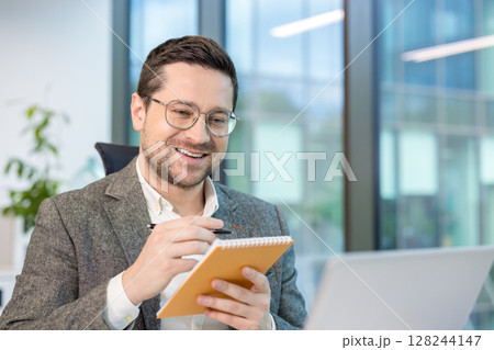Close-up photo of a young male office worker sitting in front of a laptop screen, chatting online and taking notes in a notebook. 128244147