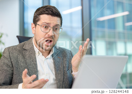 A shocked young businessman sits at a desk in the office and looks at the laptop screen in shock, spreading his arms wide. 128244148