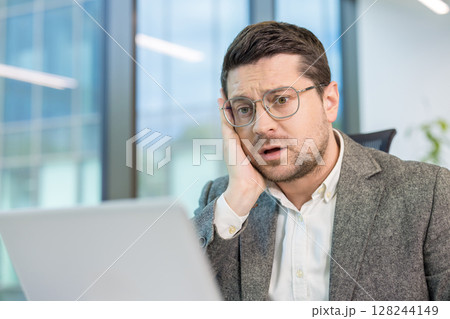 Upset young male office worker sitting at his workplace and looking disappointedly at the laptop screen. 128244149