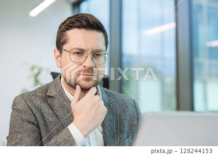 Close-up photo of a young serious man sitting in the office, looking thoughtfully at the laptop screen, solving a question and problem. 128244153