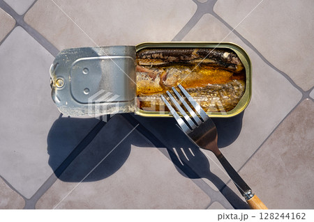 Overhead view of open sardine tin with a fork inside, casting long shadows on a tiled surface. Simple food or emergency meal. 128244162