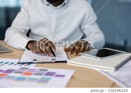 Close-up shot of a person's hands writing on a document at a desk. A tablet and notepad rest beside. 128246220