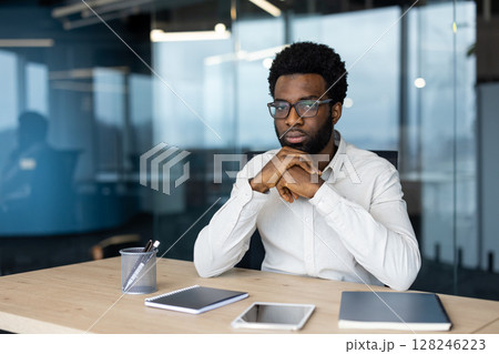 A thoughtful African-American man wearing glasses and a white shirt sits at his desk in a modern office setting. 128246223