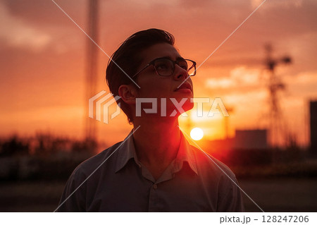 Young person with glasses gazing upwards in sunset sky. The golden hour light casts a warm glow, creating a silhouette and an atmospheric portrait. Young person with glasses gazing upwards in sunset sky. The golden hour light casts a warm glow, creating a silhouette and an atmospheric portrait. 128247206