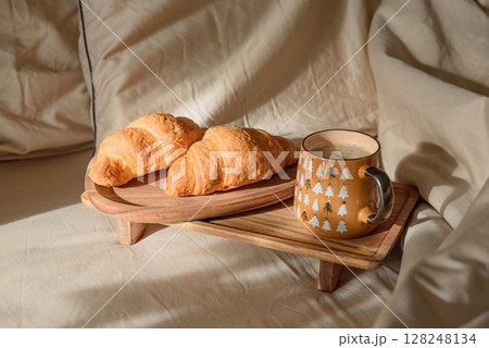 Breakfast in bed: cup of coffee and fresh croissant on wooden tray. Warm window light 128248134