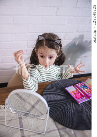 Funny little girl with sparkly cat ears experiments with toy makeup, squinting one eye while playfully dabbing pink eyeshadow in front of a mirror 128249228