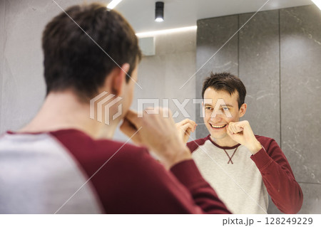 Young man flosses his teeth in front of a mirror, smiling with focus as he follows a healthy oral care routine to maintain dental hygiene and cleanliness. 128249229