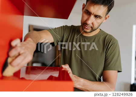 Portrait of young focused man cleaning wiping red shelf surface with sponge and detergent ensuring spotless and hygiene at home, close-up. Concept of cleanliness in domestic environment. 128249926