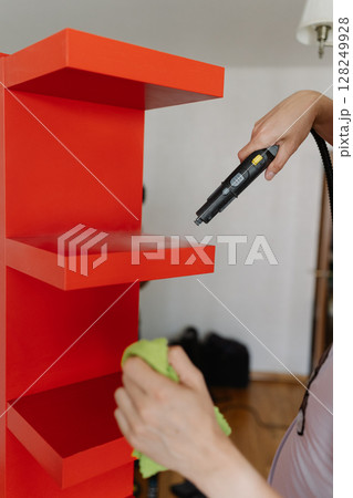 Vertical cropped shot of professional housekeeper wielding steam cleaner and green microfiber cloth sanitizing red wooden shelf, delivering comprehensive cleaning service for immaculate home interior. Vertical cropped shot of professional housekeeper wielding steam cleaner and green microfiber cloth sanitizing red wooden shelf, delivering comprehensive cleaning service for immaculate home interior. 128249928