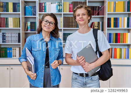 Portrait of teenage guy student with female teacher, mentor looking at camera in library Portrait of teenage guy student with female teacher, mentor looking at camera in library 128249963