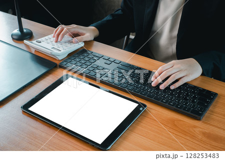 Business professional using a tablet, keyboard, and calculator at a wooden desk in a modern office setting. Business professional using a tablet, keyboard, and calculator at a wooden desk in a modern office setting. 128253483