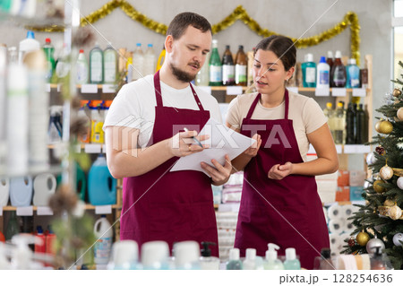 Attentive male and female sellers restock of goods, checking papers in small household store decorated for New Year celebration Attentive male and female sellers restock of goods, checking papers in small household store decorated for New Year celebration 128254636