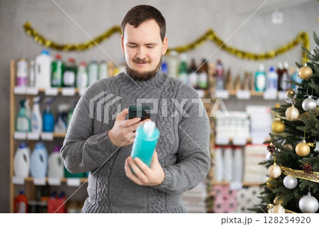 Happy handsome man choosing shampoo or shower gel and scan QR code in cosmetics store decorated for New Year holiday Happy handsome man choosing shampoo or shower gel and scan QR code in cosmetics store decorated for New Year holiday 128254920
