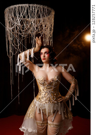 Retro-style performer in vintage costume posing on dark stage under chandelier Retro-style performer in vintage costume posing on dark stage under chandelier 128257771