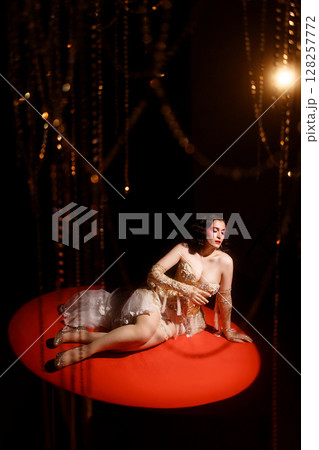Woman in vintage corset lying on red stage floor under hanging beads on dark background Woman in vintage corset lying on red stage floor under hanging beads on dark background 128257772
