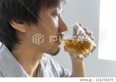 Close up shot of Asian man sitting on sofa holding glass of whiskey. Focus on the glass. Alcohol drinking concept. Close up shot of Asian man sitting on sofa holding glass of whiskey. Focus on the glass. Alcohol drinking concept. 128259274