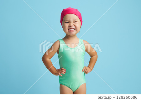 Cheerful and cute small asian girl in swim costume and cap prepares for floating in pool, dressed in swimwear, has healthy body, poses indoor against blue wall. 128260686