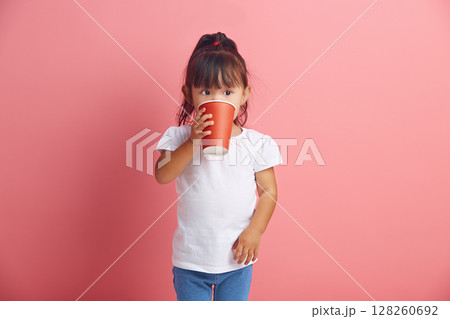 Handsome little asian girl drinking from disposable paper cup standing over pink isolated background.  128260692