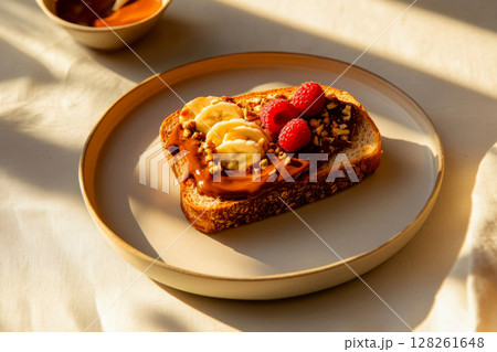 A beautifully arranged sandwich features creamy spread, fresh banana slices, vibrant raspberries, and crunchy nuts, all bathed in warm sunlight on a table 128261648