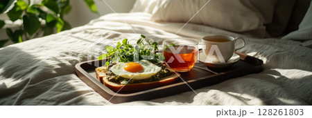 A wooden tray showcases creative toasts topped with poached eggs and vibrant herbs alongside a cup of tea. Sunlight filters through the window, adding warmth to the cozy bedroom atmosphere, banner 128261803