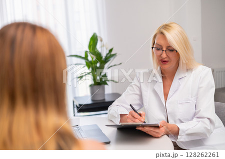 Elderly demale family physician consults young woman at home, records history, discusses recovery. Senior doctor carefully interviews female patient, makes notes, shows treatment strategy. Friendly Elderly demale family physician consults young woman at home, records history, discusses recovery. Senior doctor carefully interviews female patient, makes notes, shows treatment strategy. Friendly 128262261